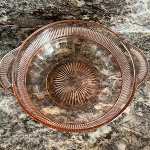 May include: A round, translucent pink glass bowl featuring two integrated side handles. The interior base displays a radiating sunburst pattern, while the wide rim is decorated with a series of vertical, parallel ridges encircling the entire edge.