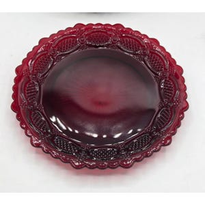 May include: A ruby red glass plate with a decorative, scalloped edge. The plate features an intricate embossed pattern around the rim and a smooth, slightly textured center. This vintage-style plate is ideal for serving or display.