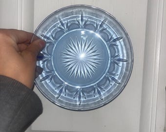 Pode incluir: Um prato de vidro azul claro com um desenho de estrela no centro. O prato tem uma borda ondulada e é segurado por uma mão. O prato tem aproximadamente 15 cm de diâmetro.