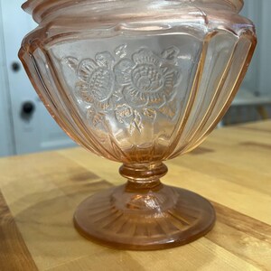Pode incluir: Uma ta&ccedil;a de vidro rosa transl&uacute;cido sobre pedestal com um padr&atilde;o floral prensado no exterior. A ta&ccedil;a tem uma borda larga e recortada, detalhes verticais canelados nas laterais e uma base circular com um design de raios de sol.