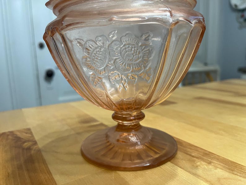 Pode incluir: Uma ta&ccedil;a de vidro rosa transl&uacute;cido sobre pedestal com um padr&atilde;o floral prensado no exterior. A ta&ccedil;a tem uma borda larga e recortada, detalhes verticais canelados nas laterais e uma base circular com um design de raios de sol.