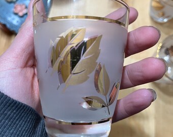 Pode incluir: Um copo de vidro fosco com design de folhas douradas. O vidro tem uma borda dourada na parte superior e inferior. O copo é segurado em uma mão. Outros copos são visíveis no fundo.