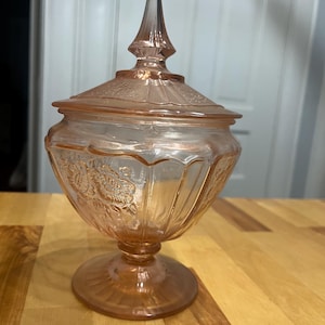 Pode incluir: Um a&ccedil;ucareiro de vidro prensado rosa com tampa a condizer. O recipiente apresenta uma base de pedestal, um remate pontiagudo na tampa e padr&otilde;es florais em relevo &agrave; volta da ta&ccedil;a. O vidro tem um tom rosa claro transl&uacute;cido em toda a pe&ccedil;a.