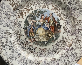 Pode incluir: Um prato decorativo vintage com um padrão floral dourado e uma cena central que retrata figuras em trajes de época. A borda do prato apresenta um padrão repetido de rosas e videiras, criando uma estética clássica e elegante.