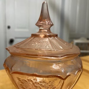 Pode incluir: Um pote de vidro rosa transl&uacute;cido com uma tampa a condizer com um remate pontiagudo. A tampa e o corpo do pote est&atilde;o decorados com padr&otilde;es florais em relevo e pain&eacute;is verticais. O vidro tem uma superf&iacute;cie lisa e reflexiva de tom rosa.