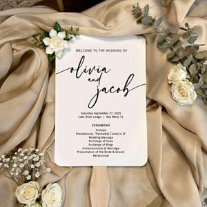 May include: A wedding program fan with the names "Olivia and Jacob" in elegant script. The program includes ceremony details and date. White flowers and greenery accent the beige background.