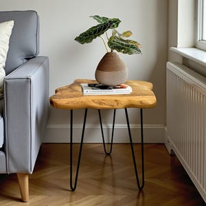 May include: A wooden side table with a natural edge and black hairpin legs. The table supports a vase with a plant and a book. The table is made of light-coloured wood and is in a living room setting.