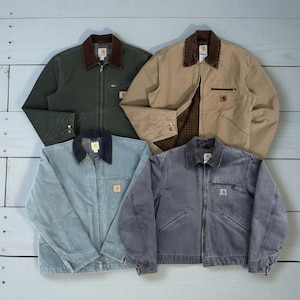 Puede incluir: Cuatro chaquetas de trabajo en varios colores: verde oscuro, beige, azul claro y gris. Cada chaqueta tiene un cuello de pana marrón, una cremallera y un pequeño parche cuadrado con el logo. Las chaquetas están sobre una superficie de madera azul claro.