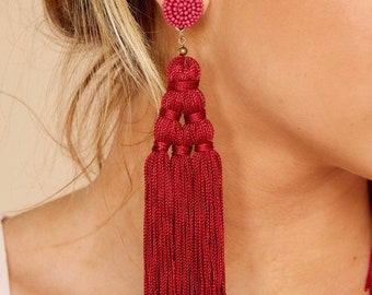 May include: A close-up of a burgundy tassel earring. The earring features a beaded stud, a series of knotted sections, and long, flowing tassels. The earring is suspended from a gold-colored hook.