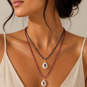 May include: Two layered necklaces with pear-shaped pendants. The top necklace features a dark blue beaded chain with a white pearl pendant and a rectangular blue gem. The bottom necklace has a red beaded chain with a white pearl pendant and a rectangular red gem.