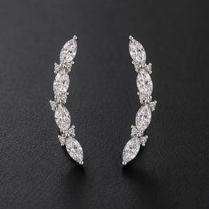 Puede incluir: Un par de pendientes trepadores plateados. Cada pendiente presenta un diseño curvo con piedras preciosas transparentes de talla marquesa y piedras redondas más pequeñas. Los pendientes están sobre un fondo oscuro.