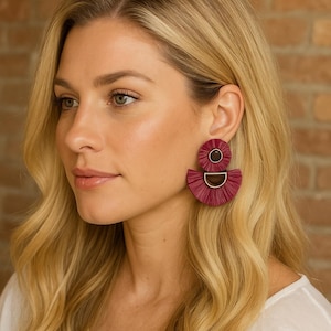 Peut inclure: Boucles d'oreilles pendantes bordeaux avec un dessus circulaire et un bas en forme d'éventail. Les boucles d'oreilles ont un accent en métal doré. La femme sur l'image a les cheveux blonds et porte une chemise blanche.