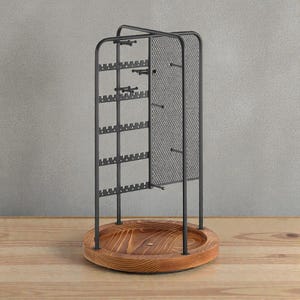 May include: A black metal jewelry organizer with a wooden base. The organizer has multiple slots for earrings and hooks for necklaces. The base is round and made of light brown wood. The background is a neutral gray.