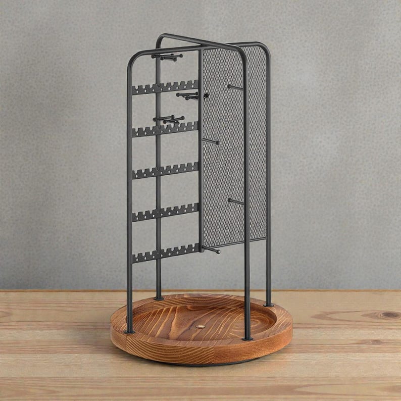 May include: A black metal jewelry organizer with a wooden base. The organizer has multiple slots for earrings and hooks for necklaces. The base is round and made of light brown wood. The background is a neutral gray.