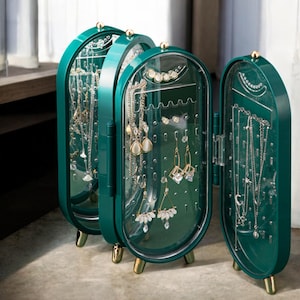May include: A teal jewelry organizer with a clear front, showcasing various necklaces and earrings. The organizer has a hinged design, gold-colored feet, and a small knob on top. The jewelry is displayed against a clear background.