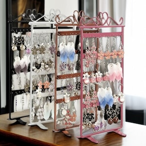 Peut inclure: Présentoirs à boucles d'oreilles noirs, blancs, bronze et roses, chacun rempli de boucles d'oreilles. Les présentoirs ont des dessus décoratifs et contiennent divers styles de boucles d'oreilles, notamment des motifs floraux, perlés et en plumes. Sur une surface en bois.