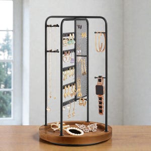 May include: A black metal jewelry organizer with a wooden base. The organizer features multiple levels for hanging necklaces, earrings, and bracelets. A watch and hoop earrings are also displayed. The jewelry stand is on a wooden table.