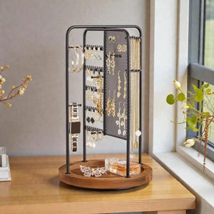 Peut inclure: Un présentoir à bijoux en métal noir avec une base en bois. Le présentoir comprend plusieurs crochets et un panneau en treillis pour suspendre boucles d'oreilles, colliers et bracelets. Une montre, du parfum et un papillon décoratif sont sur la base. Les bijoux sont dorés.