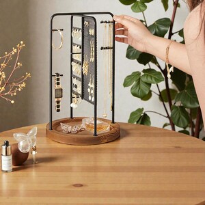 May include: A black metal jewelry organizer with a wooden base displays various earrings, necklaces, and a watch. The organizer has a rotating design and is set on a wooden table. A hand is reaching towards the jewelry.