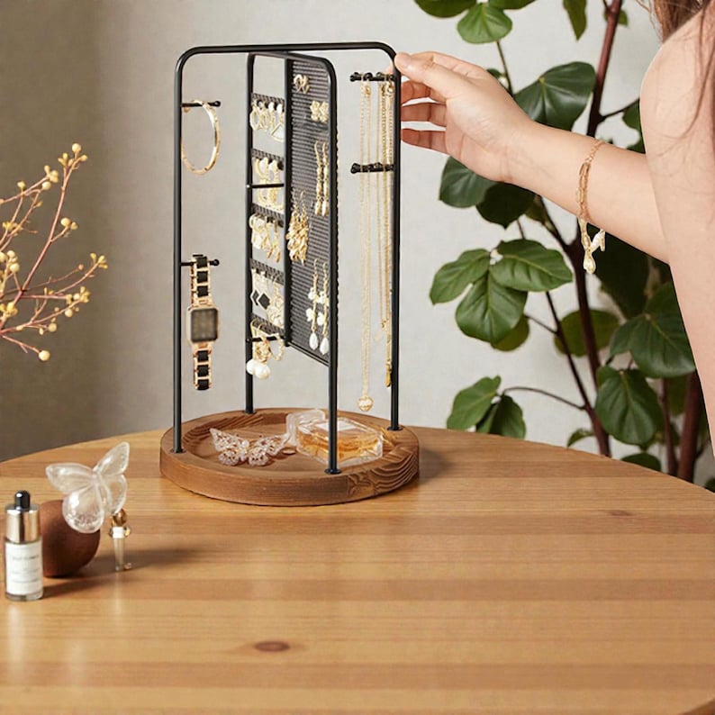 May include: A black metal jewelry organizer with a wooden base displays various earrings, necklaces, and a watch. The organizer has a rotating design and is set on a wooden table. A hand is reaching towards the jewelry.