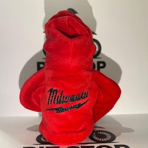 May include: A red plush hoodie featuring the word "Milwaukee" in black script on the back, with a black lightning bolt design. The hoodie has a hood and two arm-like extensions.