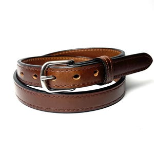 May include: A brown leather belt featuring a smooth finish, tonal stitching along the edges, and a silver-toned metal buckle. The belt is coiled into a circular shape, displaying multiple punched holes for adjustment and a single leather keeper loop.
