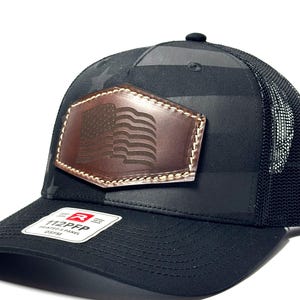May include: A black trucker hat featuring a dark gray American flag pattern on the front panels and a mesh back. A brown leather patch with an embossed American flag is stitched to the front. A sticker on the brim reads 112PFP PRINTED 5 PANEL OSFM.