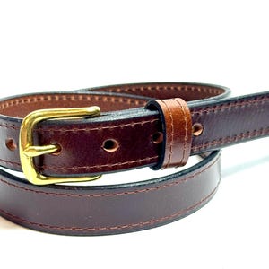 May include: A dark brown leather belt featuring a smooth finish, visible tonal stitching along the edges, and a polished gold-tone metal buckle. The belt is coiled, showing multiple punched holes for adjustment and a matching leather keeper loop.