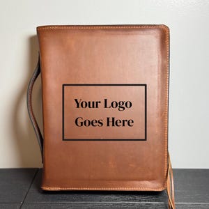 May include: A brown leather portfolio with a handle and strap. The text "Your Logo Goes Here" is displayed in a black rectangle on the front. The portfolio is rectangular and appears to be new.