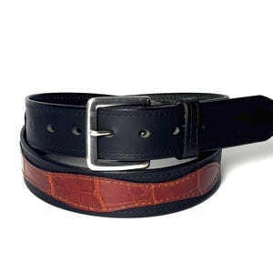May include: A black leather belt featuring a silver-toned metal buckle and a contrasting brown textured inlay with a reptilian-style pattern. The belt is coiled, showing a smooth black finish on the upper strap and a distinct brown section.