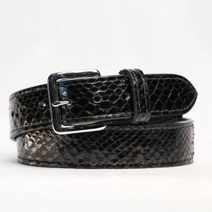 May include: A black belt with a snakeskin pattern. The belt has a silver-colored square buckle and a matching belt loop. The belt is made of a shiny, textured material.