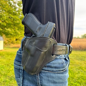May include: A black handgun in a black leather holster attached to a belt. The belt is worn with blue jeans. The holster is designed to be worn on the hip. The background shows a field and a cloudy sky.