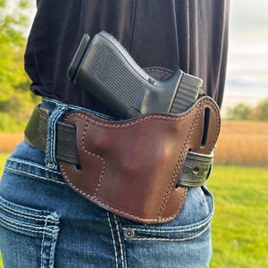 May include: A brown leather holster with a black handgun secured on a person's waist. The holster is attached to a black belt worn with blue jeans. The background shows a field and sky.