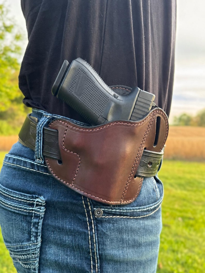 May include: A brown leather holster with a black handgun secured on a person's waist. The holster is attached to a black belt worn with blue jeans. The background shows a field and sky.