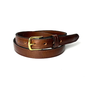 May include: A medium brown leather belt featuring a polished gold-tone metal buckle and a single matching leather keeper loop. The belt has visible stitching along the edges and multiple punched holes for adjustment, coiled into a circular shape.
