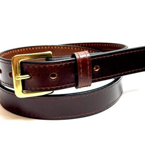 May include: A dark brown leather belt featuring a polished gold-tone metal buckle and a matching leather keeper loop. The belt has visible stitching along the edges and multiple punched holes for adjustment, coiled into a circular shape.