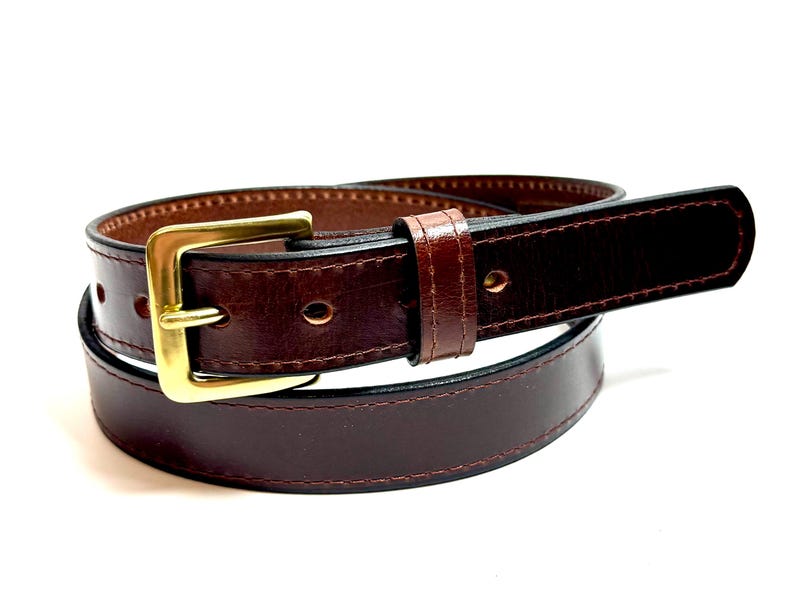 May include: A dark brown leather belt featuring a polished gold-tone metal buckle and a matching leather keeper loop. The belt has visible stitching along the edges and multiple punched holes for adjustment, coiled into a circular shape.