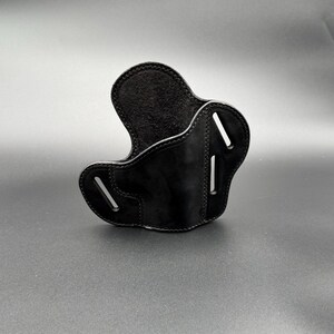 May include: A black leather holster with a curved design. The holster has a smooth, glossy finish and visible stitching along the edges. It features two vertical slots and a curved opening for a firearm.