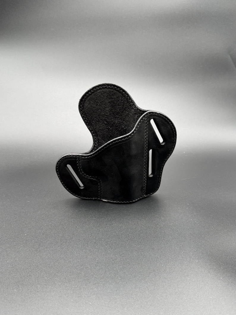 May include: A black leather holster with a curved design. The holster has a smooth, glossy finish and visible stitching along the edges. It features two vertical slots and a curved opening for a firearm.