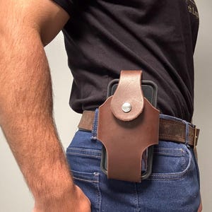 May include: A brown leather phone holster attached to a belt. The holster has a snap closure and is designed to hold a smartphone. The person is wearing a black t-shirt and blue jeans.