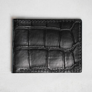May include: A black, rectangular wallet with a textured, crocodile-like pattern. The wallet is made of leather and has visible stitching around the edges. The wallet is a minimalist design, suitable for carrying cards and cash.