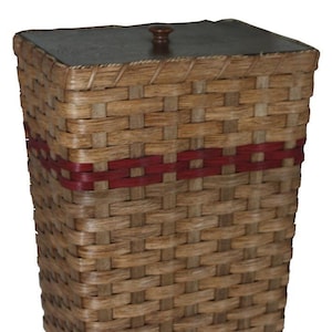 May include: A tall, rectangular woven basket with a natural light brown finish and a single horizontal band of dark red woven material. The basket features a flat, dark brown wooden lid with a small, centered wooden knob handle on top.