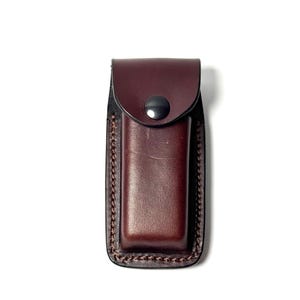 May include: A brown leather sheath with a black snap closure. The sheath is rectangular with a stitched border and is designed to hold a small tool or knife. The leather has a smooth finish.
