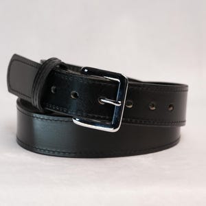 May include: A black leather belt with a silver-colored buckle. The belt is rolled up, showcasing its smooth texture and stitching details. The buckle is square and shiny.