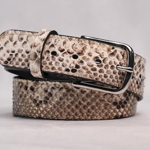 May include: A coiled belt with a snakeskin pattern in shades of beige and brown. The belt features a silver-colored rectangular buckle and a matching belt loop. The leather belt is a fashion accessory.