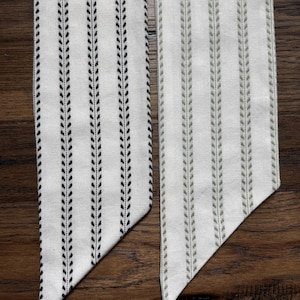 May include: Two long, narrow fabric strips with pointed ends. One strip features a white background with vertical black leaf-patterned stripes, while the other features a white background with vertical sage green leaf-patterned stripes.