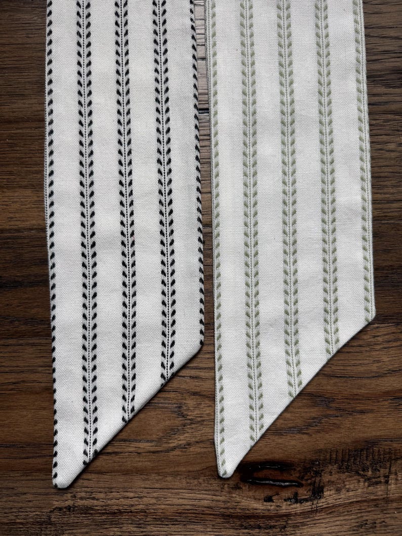 May include: Two long, narrow fabric strips with pointed ends. One strip features a white background with vertical black leaf-patterned stripes, while the other features a white background with vertical sage green leaf-patterned stripes.