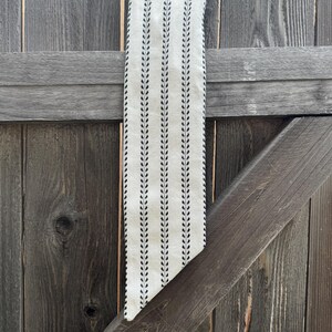 May include: A long, narrow fabric scarf in an off-white cream color featuring a repeating vertical pattern of thin black lines and small, delicate black leaf-like motifs arranged in a symmetrical, linear design with a pointed, angled end.