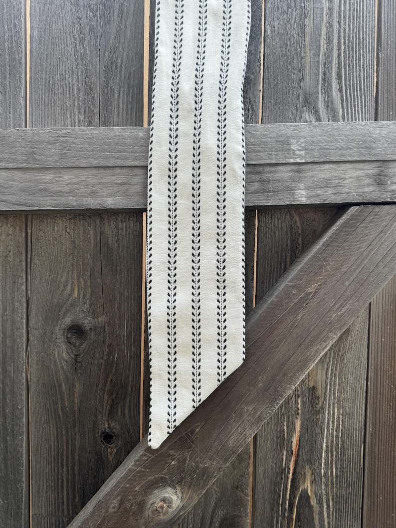 May include: A long, narrow fabric scarf in an off-white cream color featuring a repeating vertical pattern of thin black lines and small, delicate black leaf-like motifs arranged in a symmetrical, linear design with a pointed, angled end.