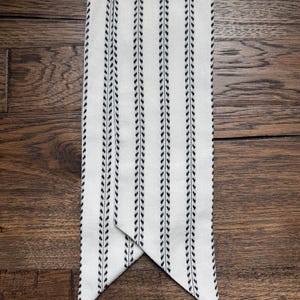 May include: A long, rectangular fabric scarf in an off-white color featuring vertical stripes of black, repeating leaf-like patterns. The scarf has pointed, triangular ends that overlap slightly, creating a clean and symmetrical appearance.